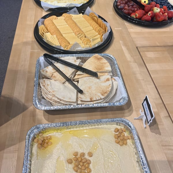 A buffet spread with crackers, pita bread, hummus, and a fruit platter on a wooden table.