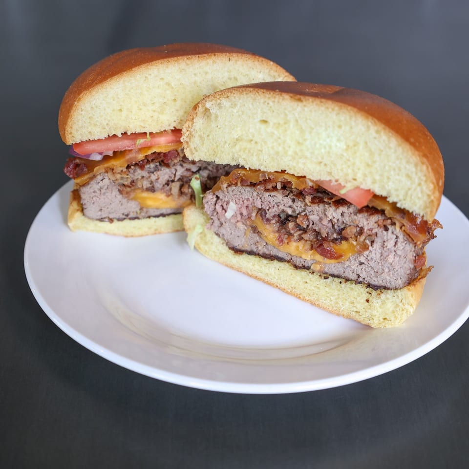 Two halves of a Burger Shack burger with cheese, bacon, and tomato on a white plate.