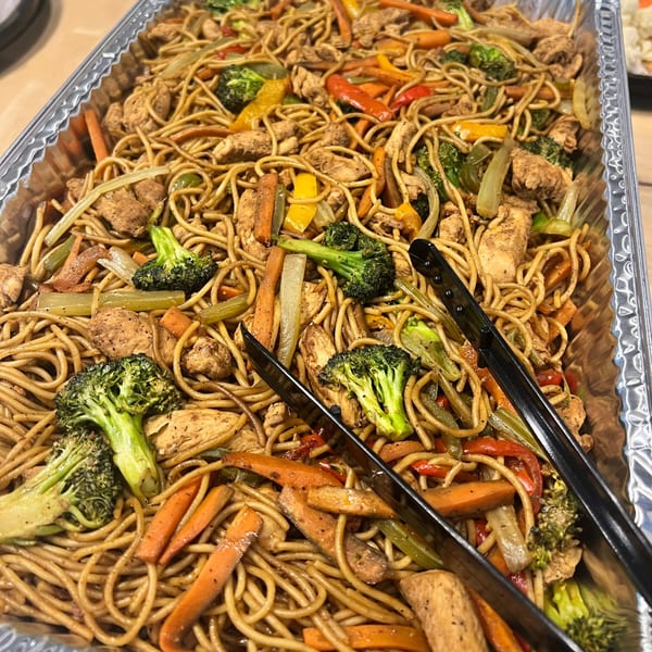Large catering tray of chicken lo mein with broccoli, carrots, and bell peppers.