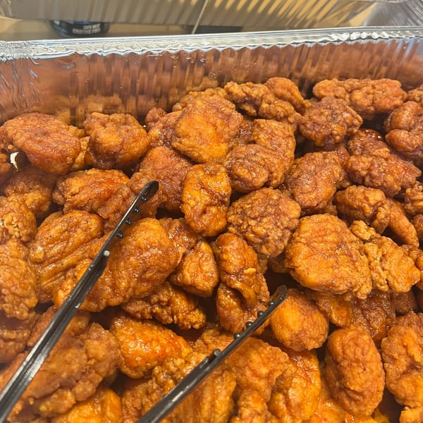 Close-up of crispy, orange-glazed boneless chicken wings in a foil tray with serving tongs.