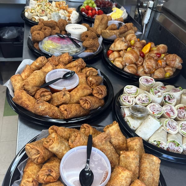 Multiple catering trays filled with various appetizers, including egg rolls, wraps, falafel, and croissants.
