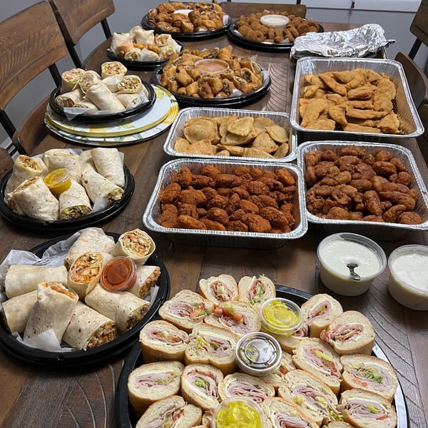 A catering spread of chicken tenders, wraps, subs, and fried appetizers on a wooden table.