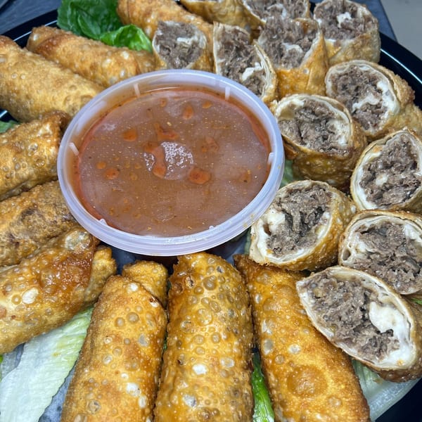 Crispy cheesesteak egg rolls, some cut open to show filling, with a side of dipping sauce.