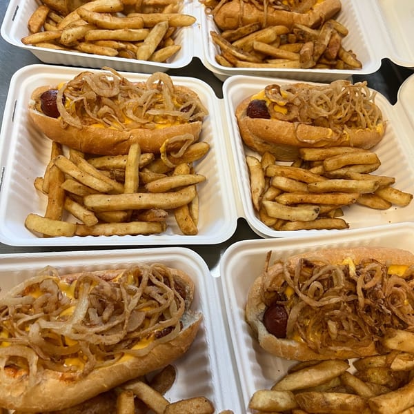 Multiple hot dogs with cheese and crispy fried onions, served with french fries in white takeout containers.